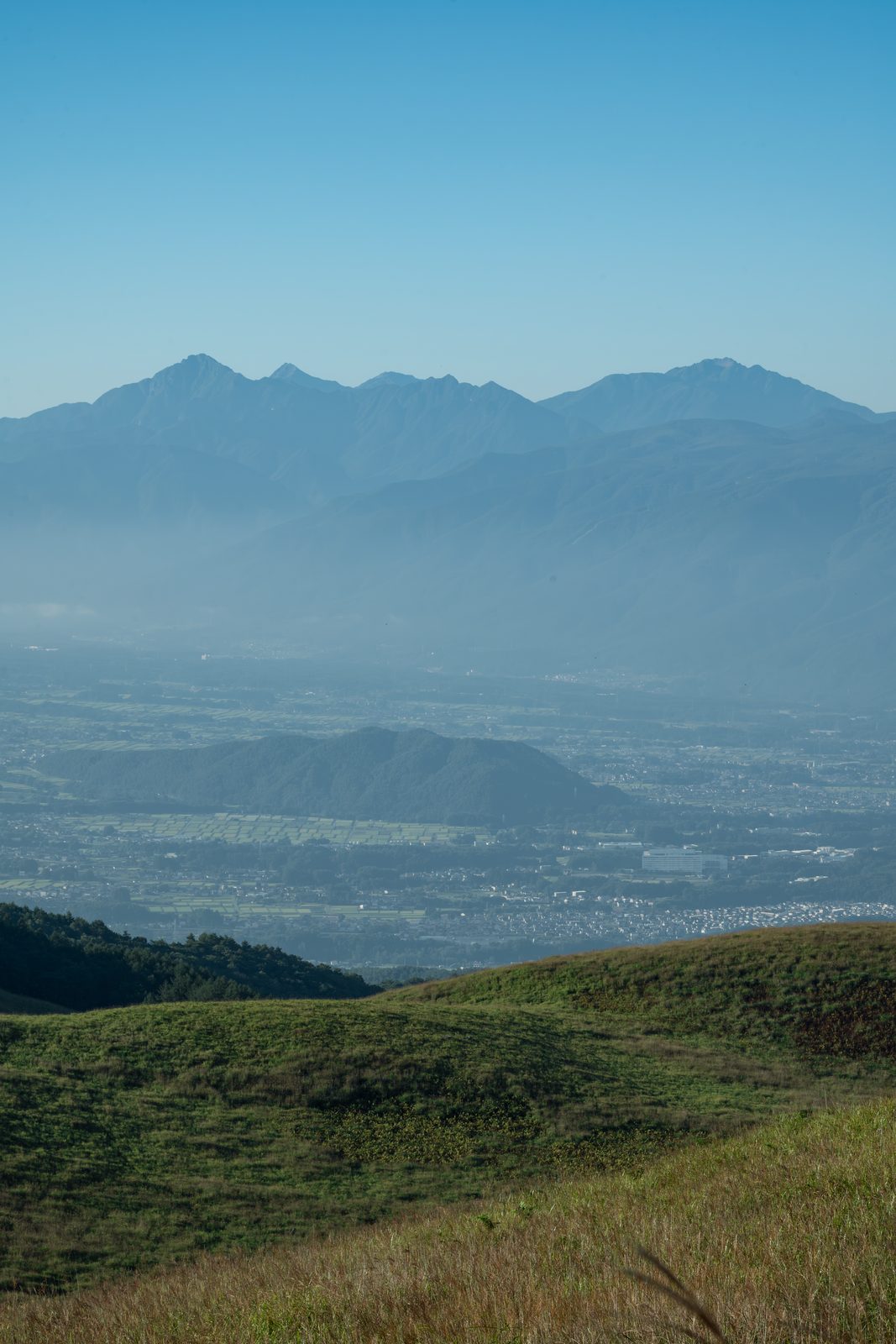 草地の斜面の先に広がる平野と、その奥に連なる山並み